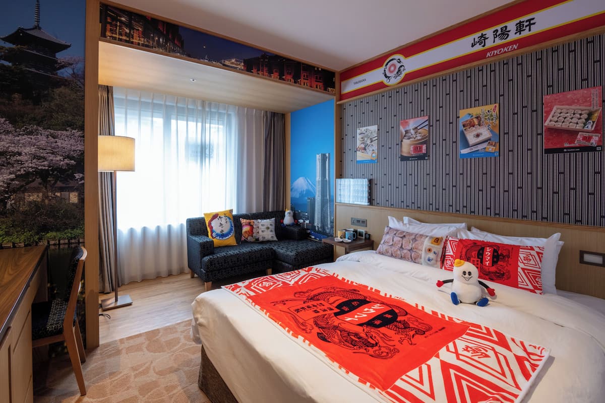 A hotel room with a bed, a desk, a chair, a nightstand, and a wall with posters.