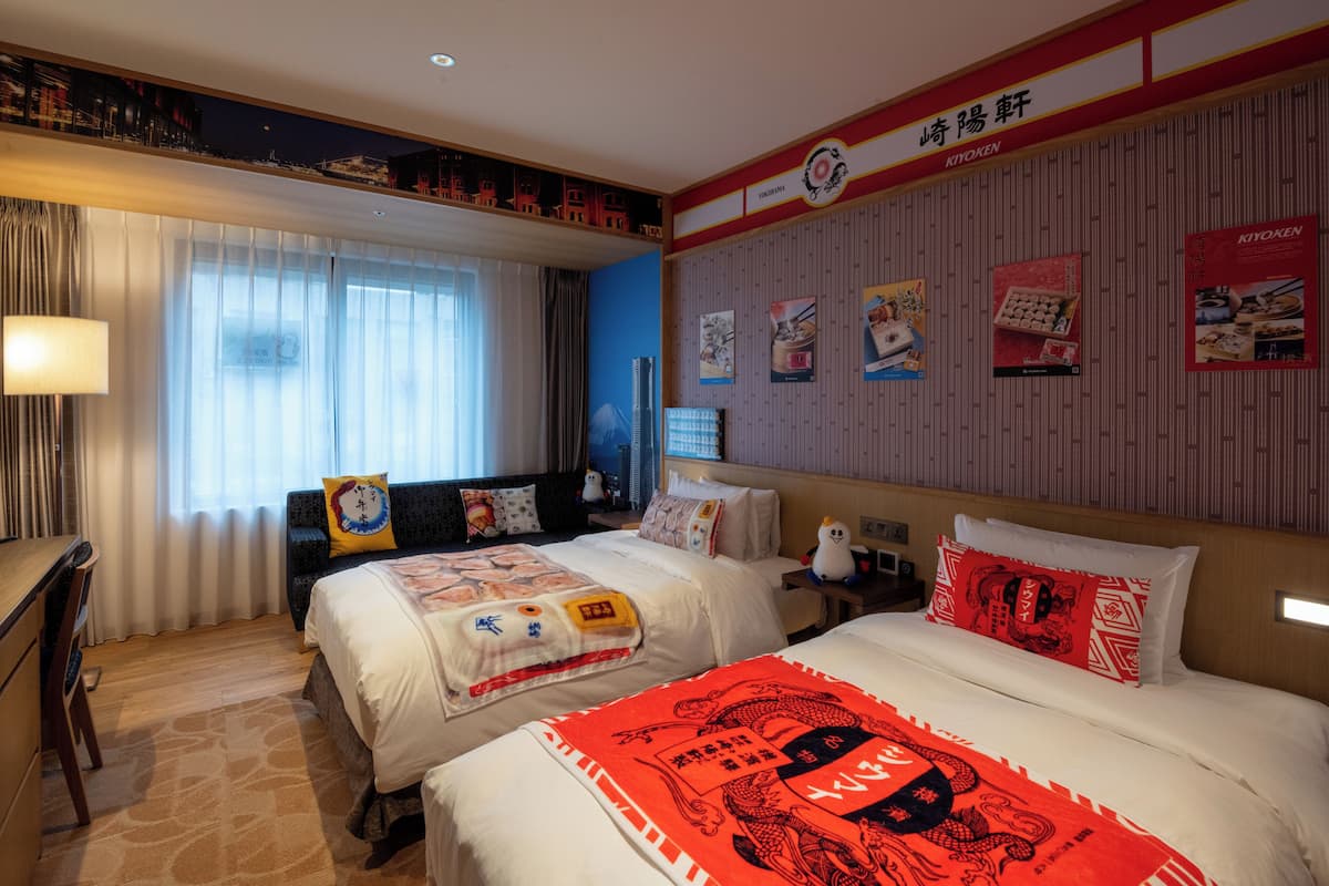 A hotel room with two beds, a desk, a chair, a nightstand, and a wall with posters.