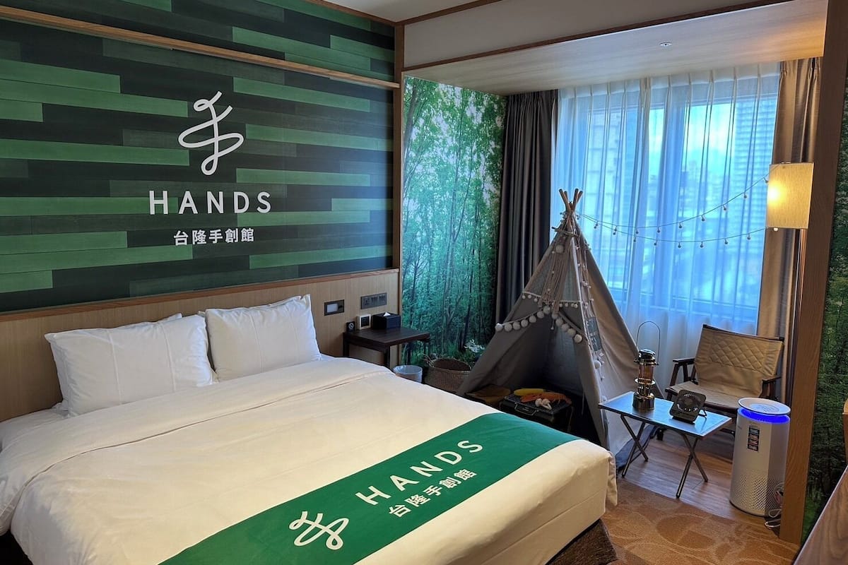 A hotel room with a bed, a green wall with the 'HANDS' logo, a teepee, and a view of trees through the window.