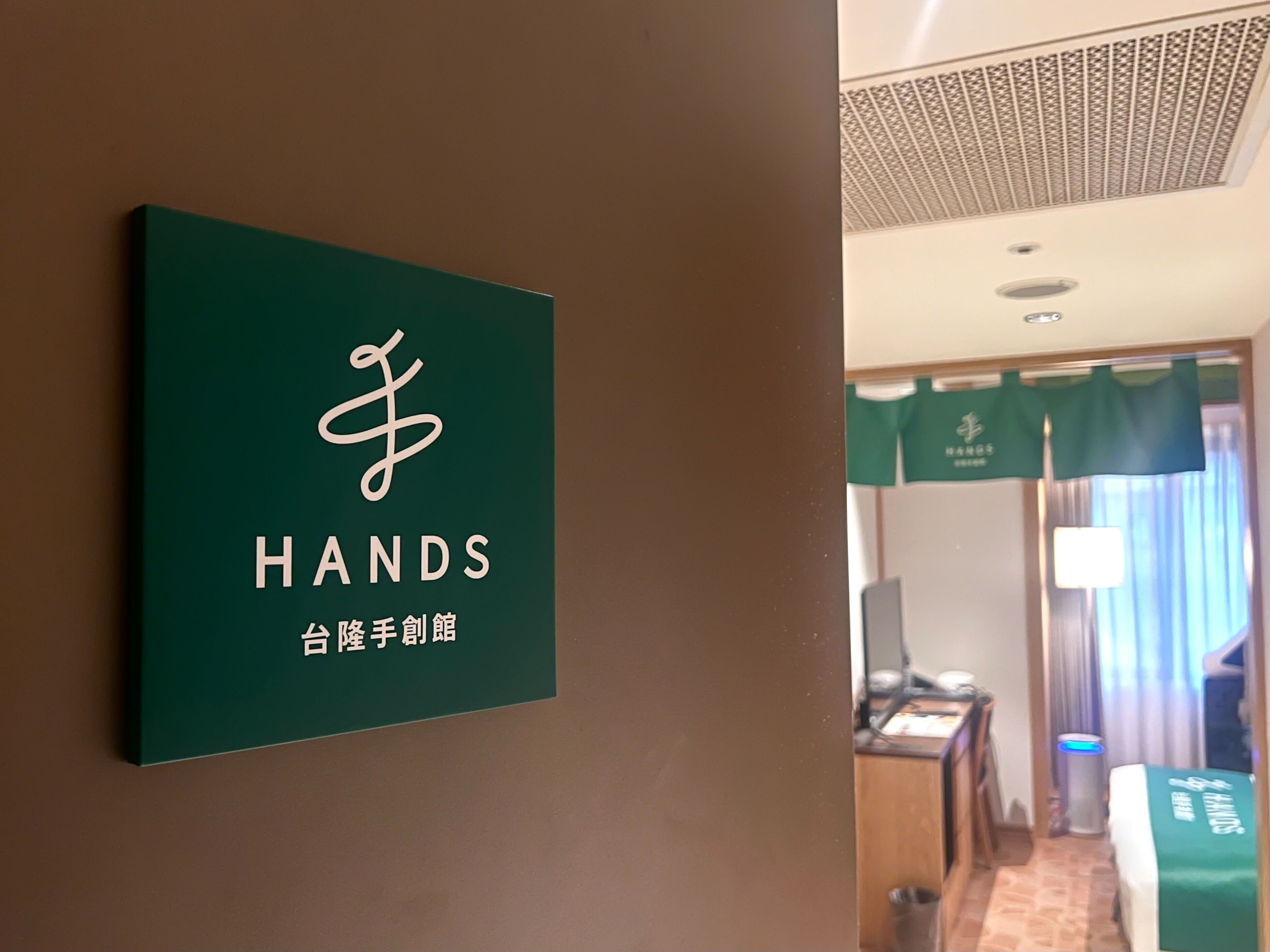 A hotel room with a green sign on the wall, a bed with a green cover, a desk, and a chair.