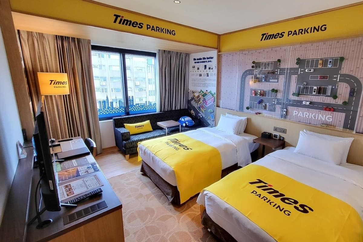 A hotel room with two beds, a desk, a TV, a window with curtains, and a Times Parking sign on the wall.