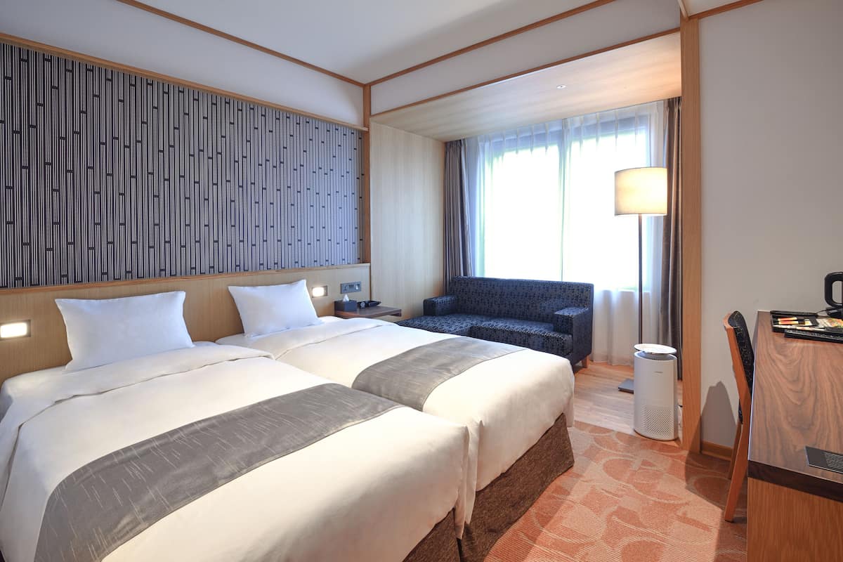 A hotel room with two beds, a desk, a chair, a lamp, and a large window with curtains.