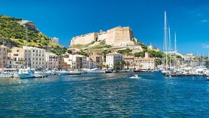 Un pueblo costero con una fortaleza histórica en una colina, barcos amarrados a lo largo del muelle y edificios con balcones.
