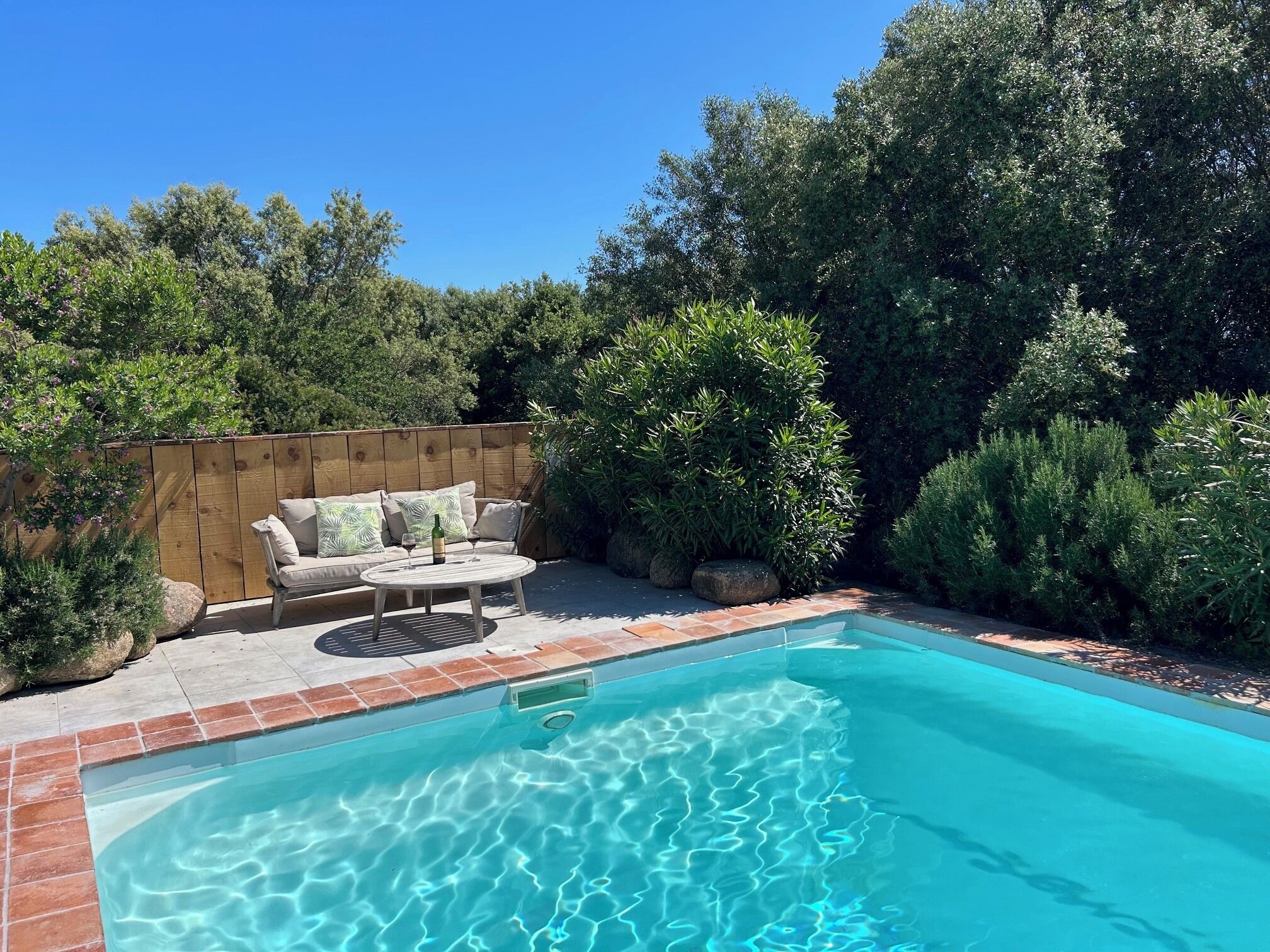 A swimming pool with a wooden deck, a white outdoor sofa, and surrounding greenery.