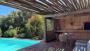 Un área junto a la piscina con un porche de madera, muebles de mimbre y una zona de cocina con televisor.