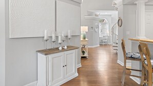 A hallway with a wooden floor, white walls, and a white cabinet with candles. There is a chair with a patterned cushion and a small table with a lamp.