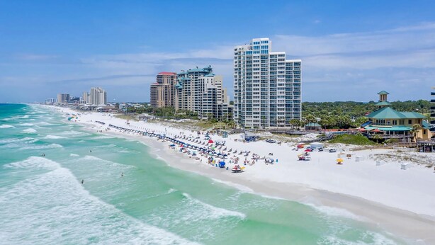 A beach with a long pier, several high-rise buildings, and a clear blue sky.