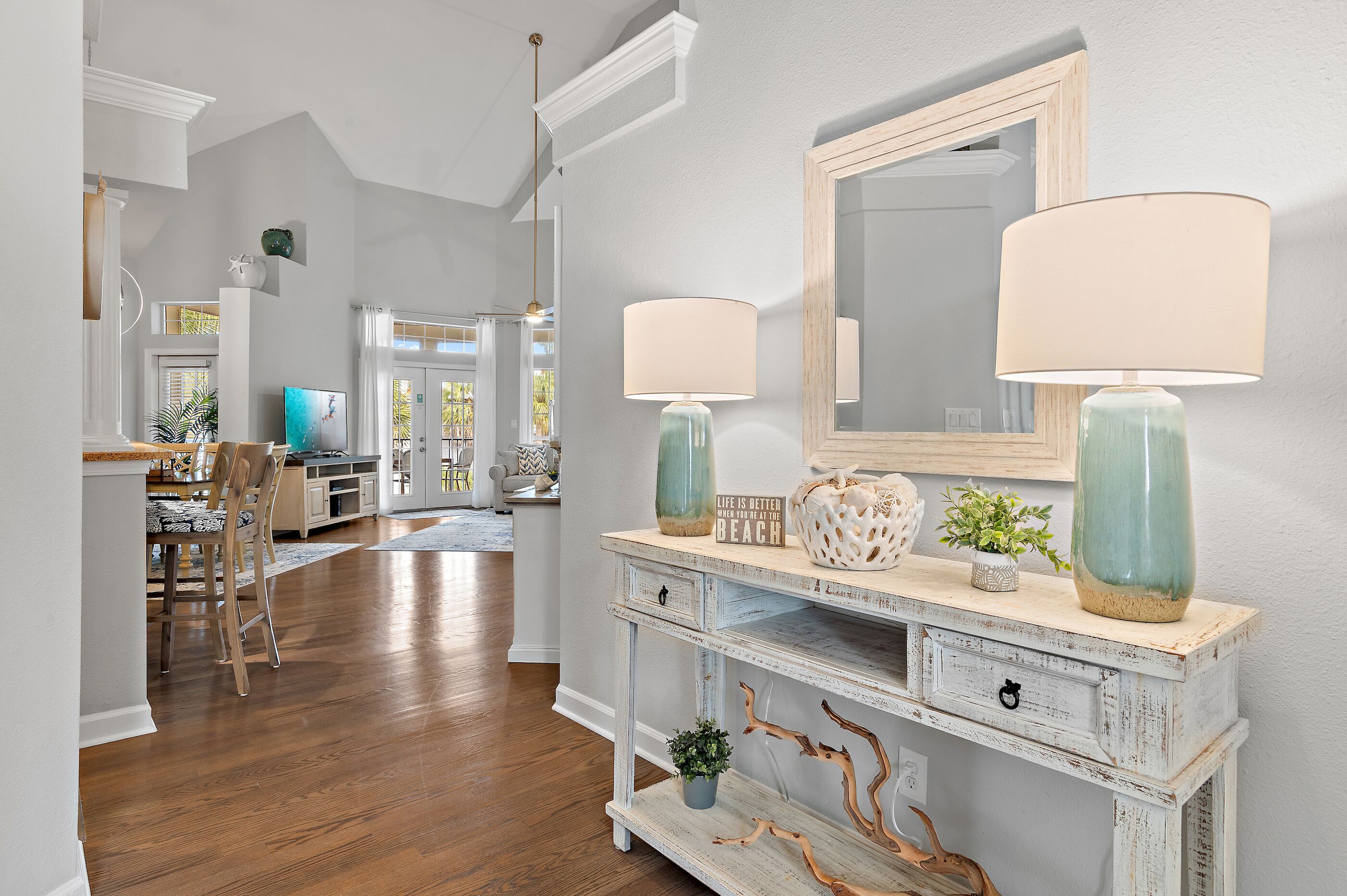 A well-lit living room with a wooden floor, a white console table with decorative items, and a large mirror above it.