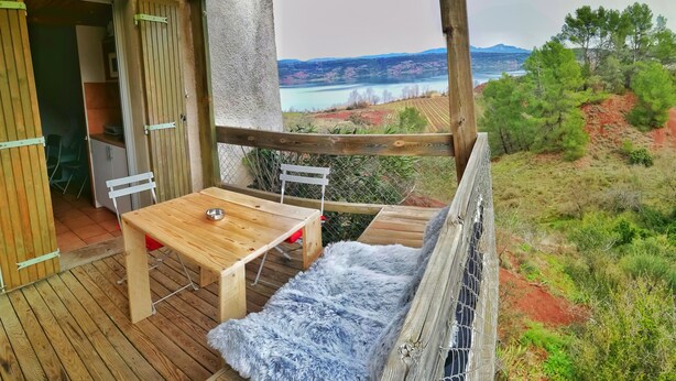 Una terraza de madera con una mesa y sillas, un banco acolchado y vistas a un lago y montañas.