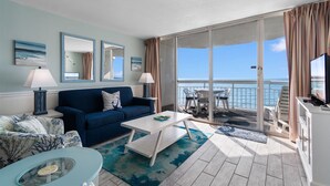 A living room with a blue sofa, a coffee table, and a view of the ocean through large sliding glass doors. - Garden City Beach Oceanfront Condo 609 | Rising Tides Properties Rental (Murrells Inlet)