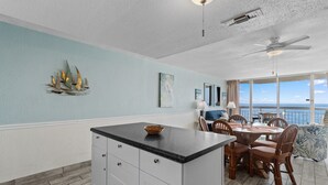 A kitchen with a central island, dining area with chairs, a ceiling fan, and a view of the ocean. - Garden City Beach Oceanfront Condo 609 | Rising Tides Properties Rental (Murrells Inlet)