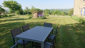 Mesa com cadeiras em área externa gramada, com um edifício de pedra e árvores ao fundo.