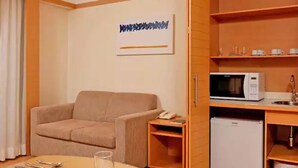 A compact hotel room with a beige sofa, a small round table with a glass, a microwave, and a mini-fridge. - Riema Paulista Classic Flat Service (São Paulo)