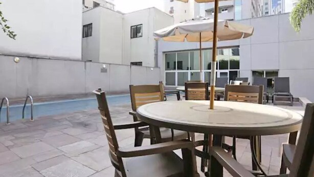 A poolside area with a round table and chairs, a pool, and a building in the background. - Riema Paulista Classic Flat Service (São Paulo)