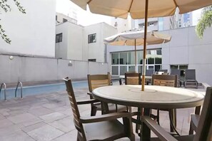 A poolside area with a round table and chairs, a pool, and a building in the background.