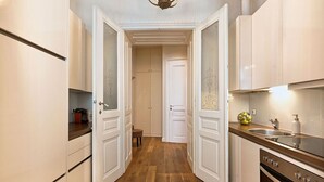 Une cuisine étroite avec des meubles blancs, un plancher en bois et une porte visible donnant sur une autre pièce.