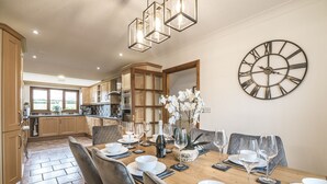 A dining area with a wooden table set for six, surrounded by chairs. A large wall clock is visible, and the room has a modern kitchen in the background. - French Fields - 4 Bedroom - St Florence (Tenby)