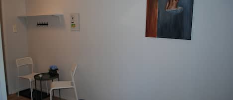 A room with a bed, a chair, a small table with a vase, a coat rack, and a painting on the wall.