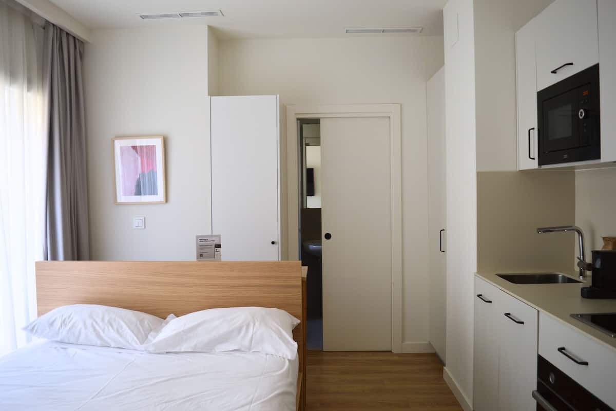Une chambre d’hôtel comprenant un lit, une kitchenette, une porte donnant accès à une salle de bain et un tableau encadré au mur.