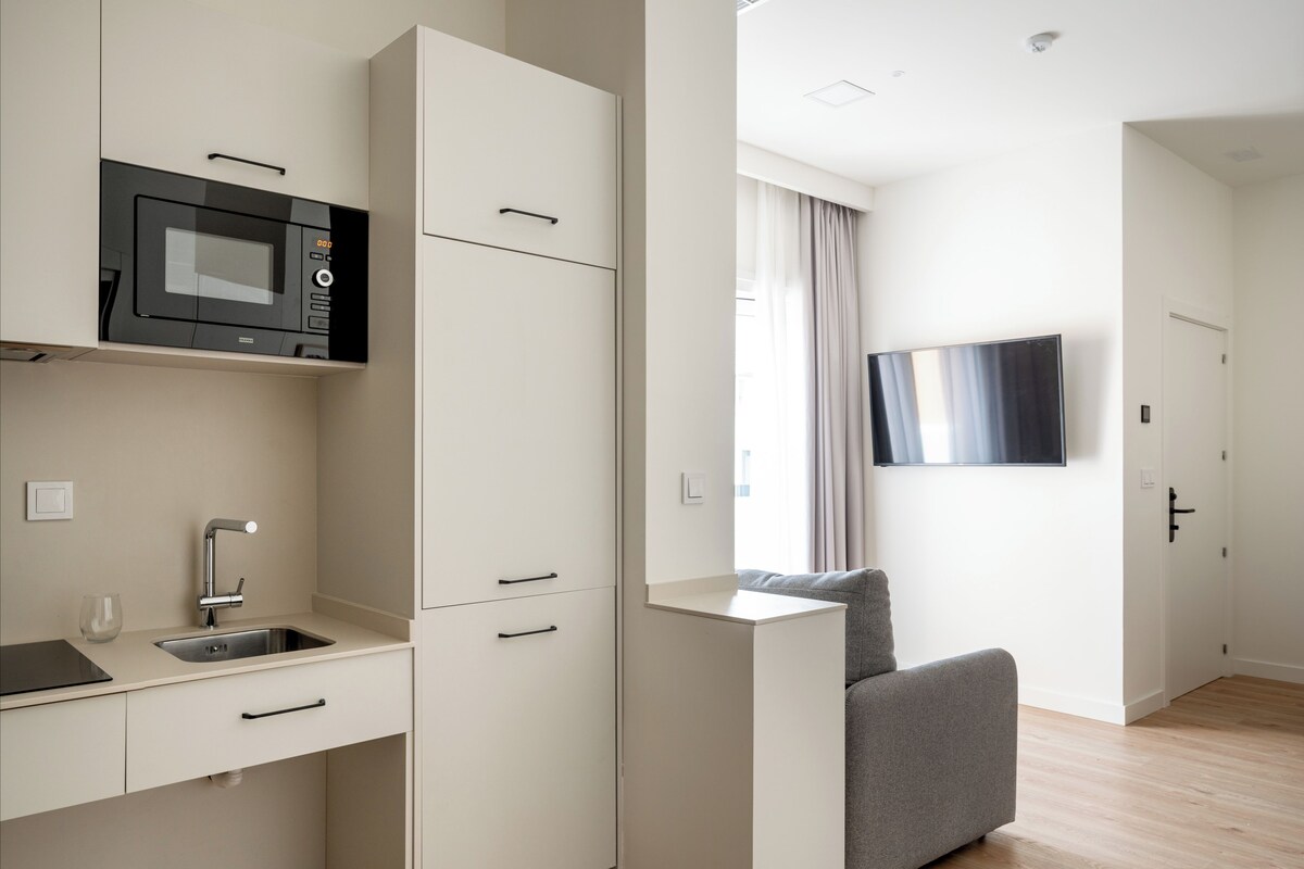 Une cuisine moderne et compacte, dotée d’un micro-ondes encastré, d’un évier et d’un petit plan de travail. On y trouve également un canapé gris et un téléviseur à écran plat fixé au mur.