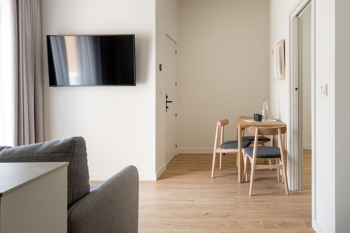 Une chambre moderne et minimaliste, dotée d’un téléviseur à écran plat fixé au mur, d’un canapé gris, d’une table à manger en bois avec deux chaises et d’un plancher en bois.
