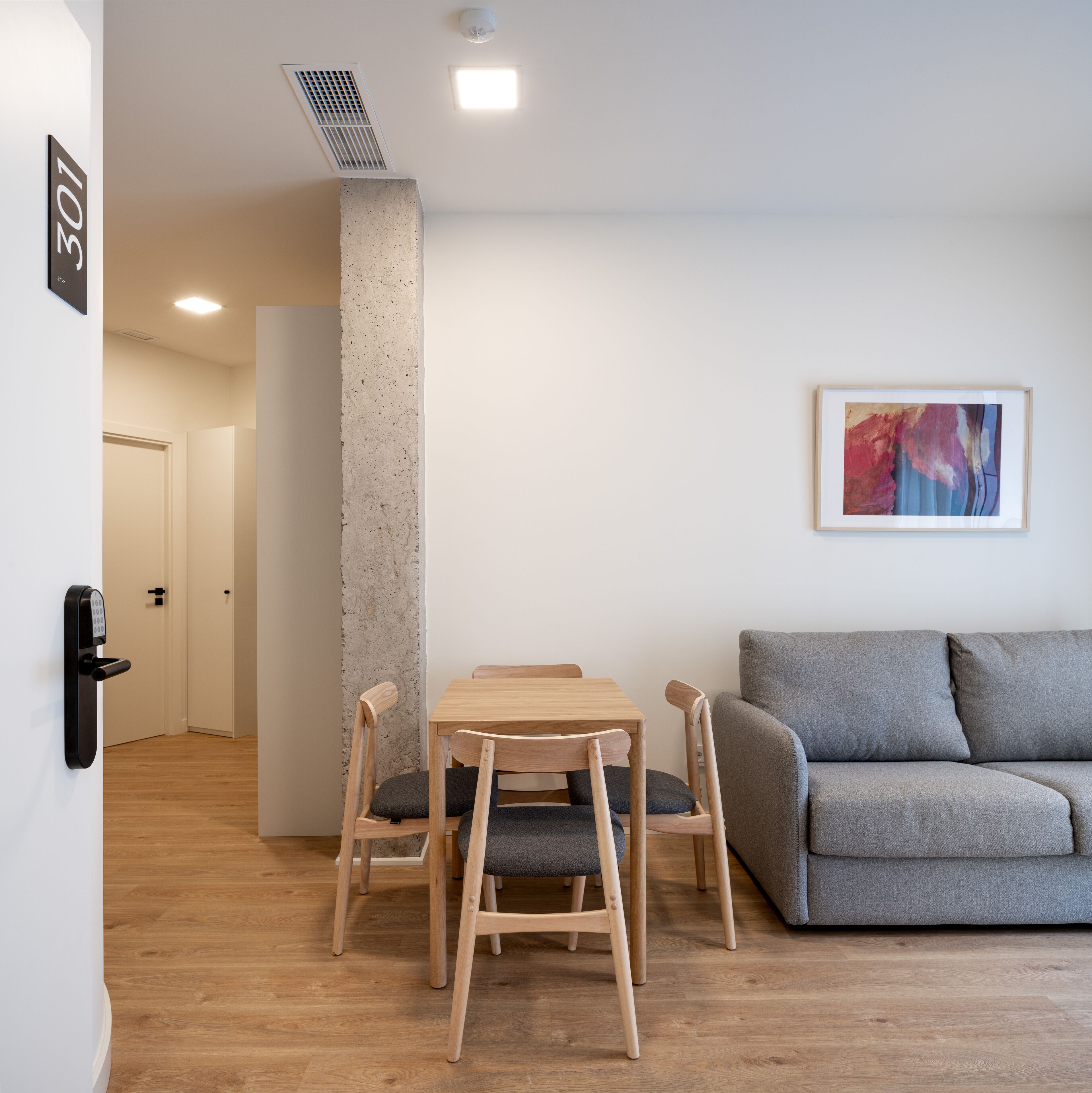 Una sala de estar moderna con un sofá gris, una mesa de comedor de madera con sillas y un pilar de concreto.