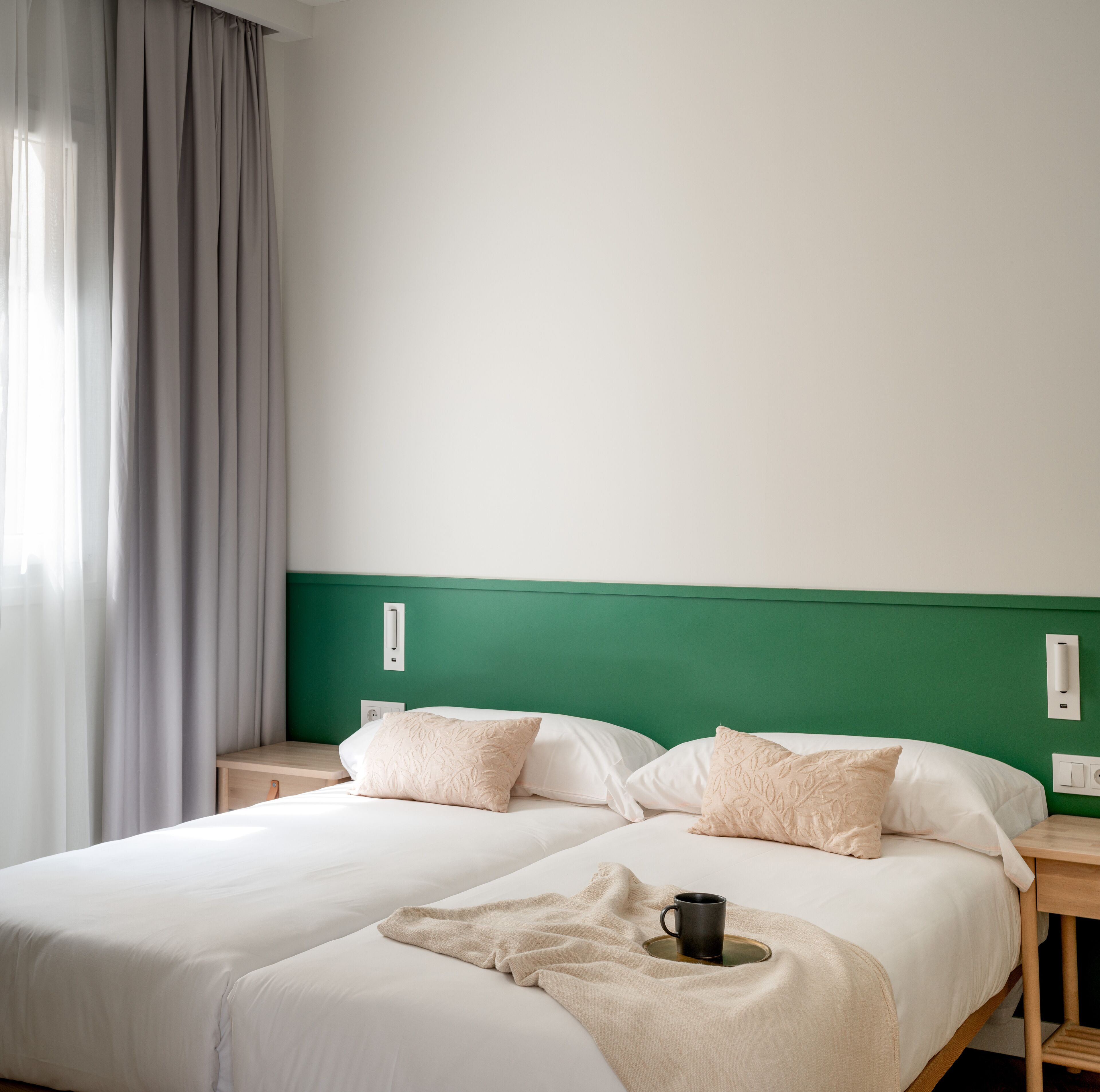 Ein Hotelzimmer mit zwei Betten, einem grünen Kopfteil und einem Fenster mit Vorhängen.