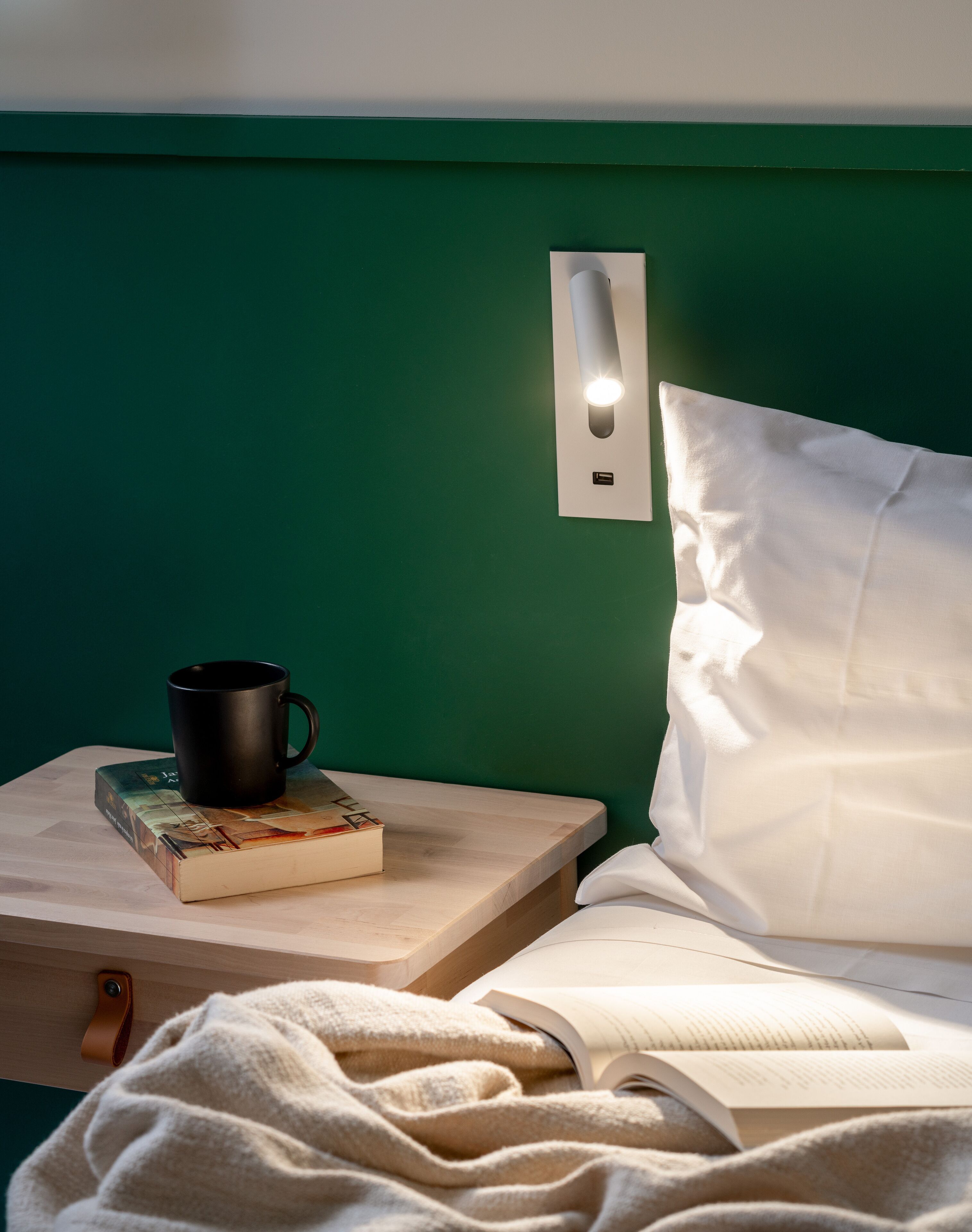 Una cama bien hecha con almohadas y edredón blancos, una mesita de noche con un libro y una taza negra, y una lámpara de pared.
