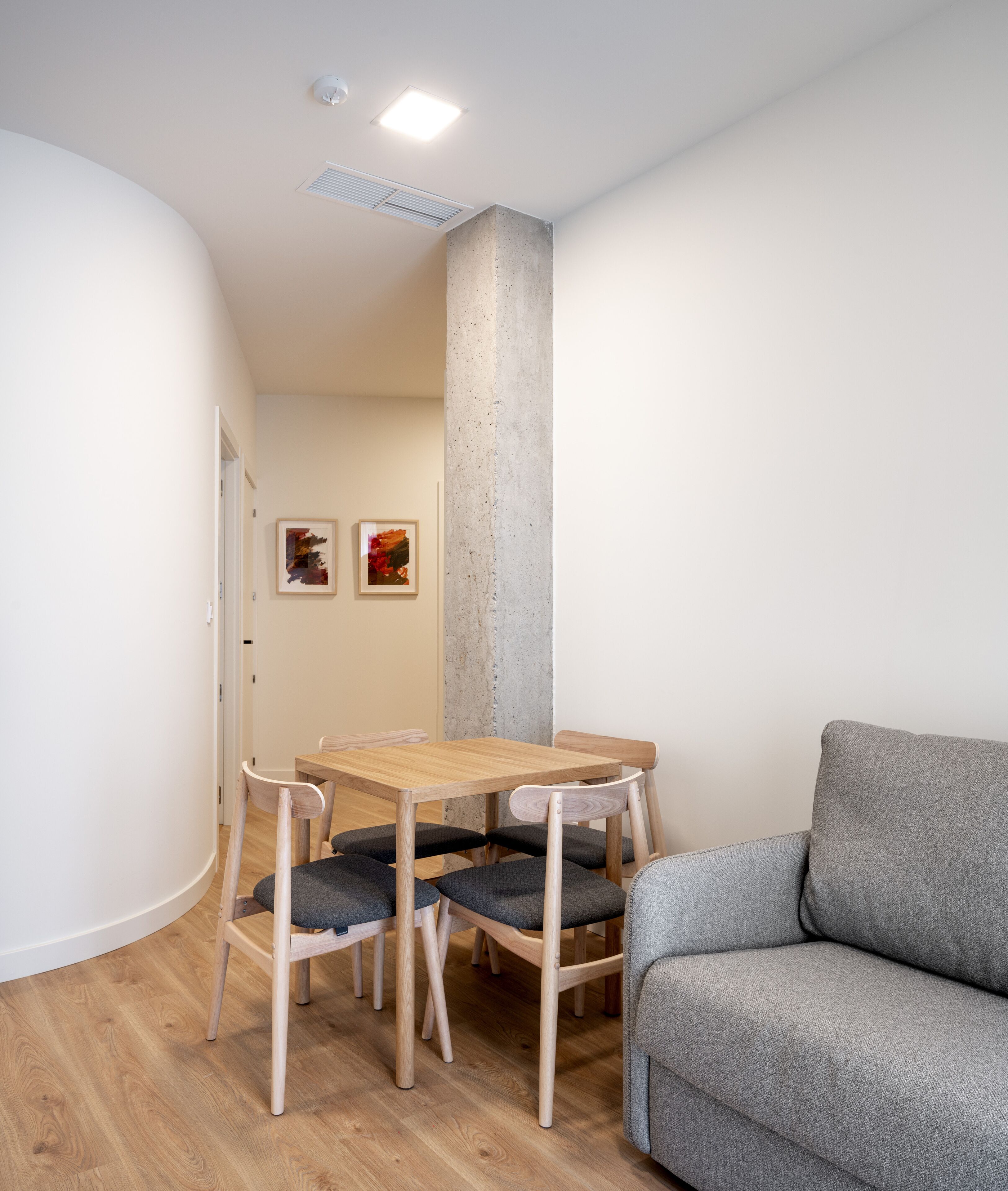 Una stanza piccola e moderna con un tavolo da pranzo in legno e quattro sedie, un divano grigio e due quadri astratti alle pareti.