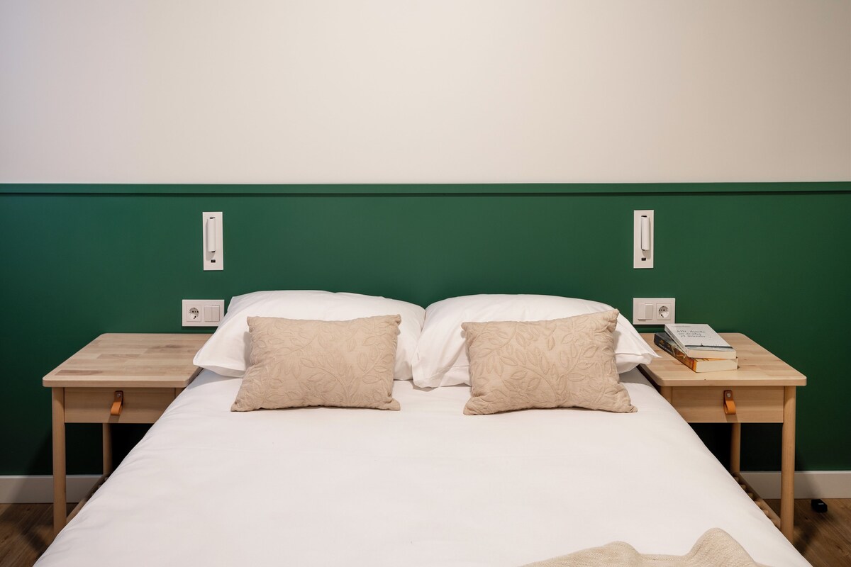 Una cama con sábanas blancas, dos almohadas color beige y una mesita de noche de madera con un libro y un teléfono.