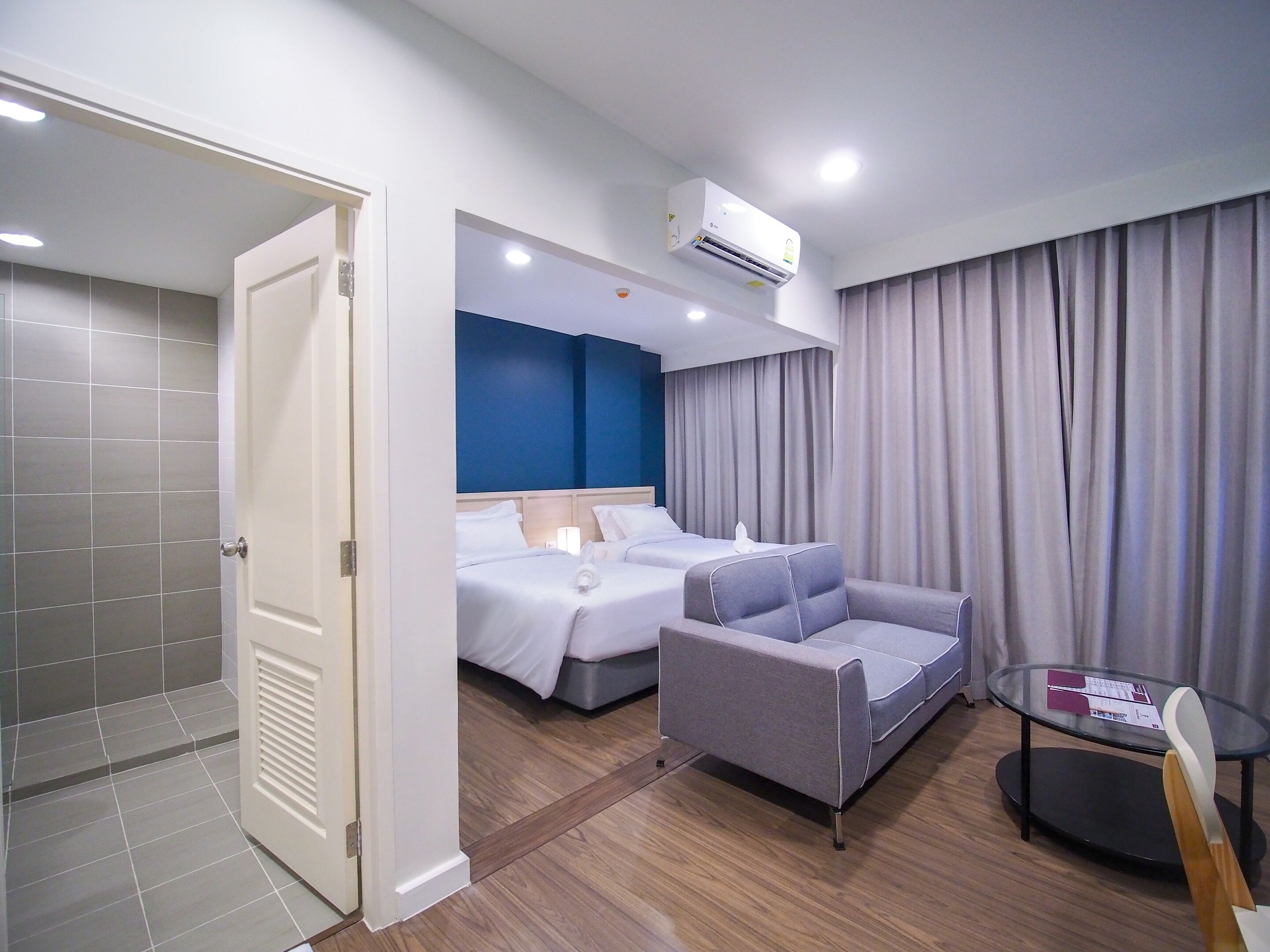 A hotel room with a bed, a sofa, a small table, and an air conditioning unit.