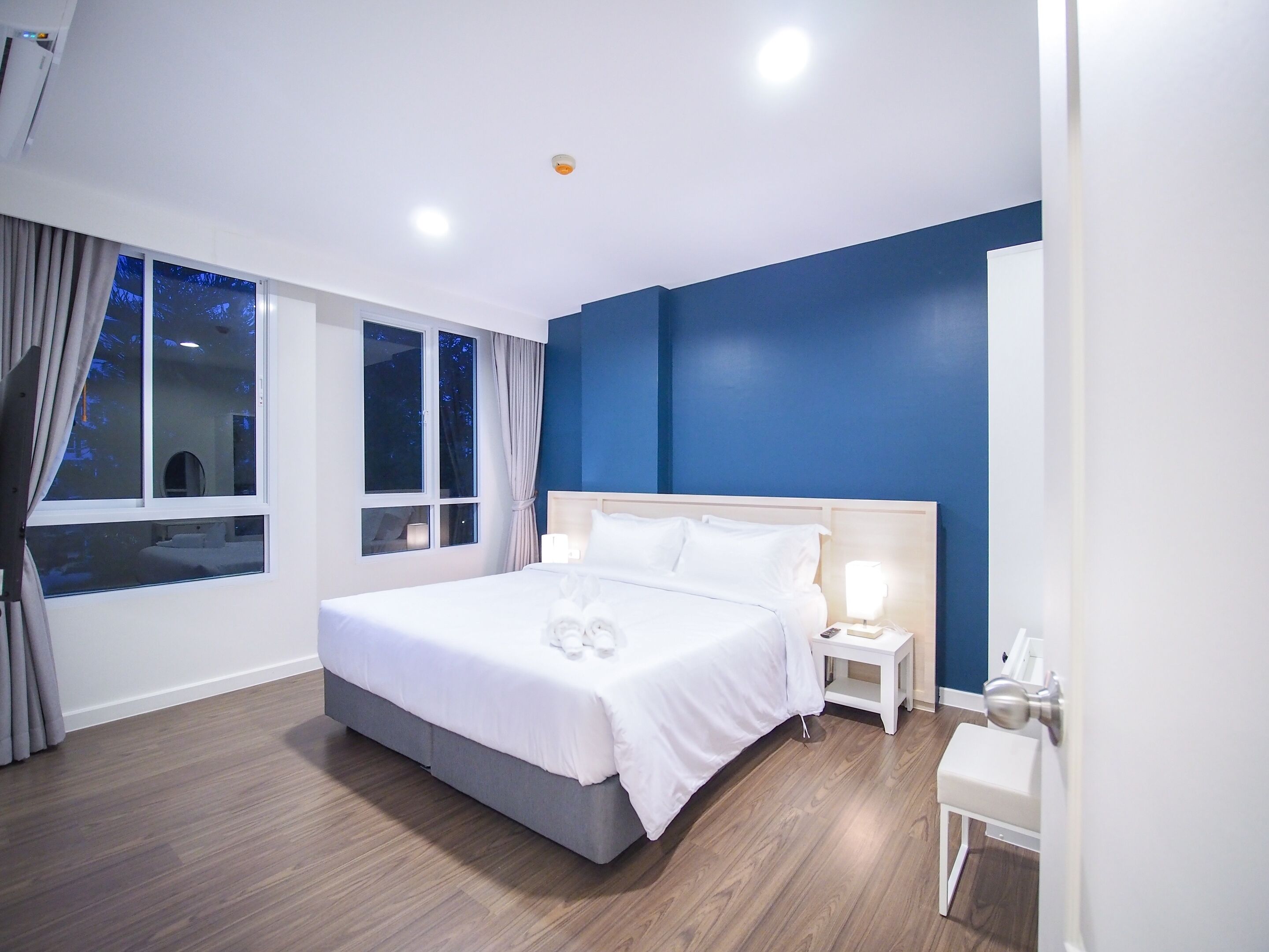 A modern hotel room with a large bed, bedside tables, a nightstand, a small desk, and a window with curtains.
