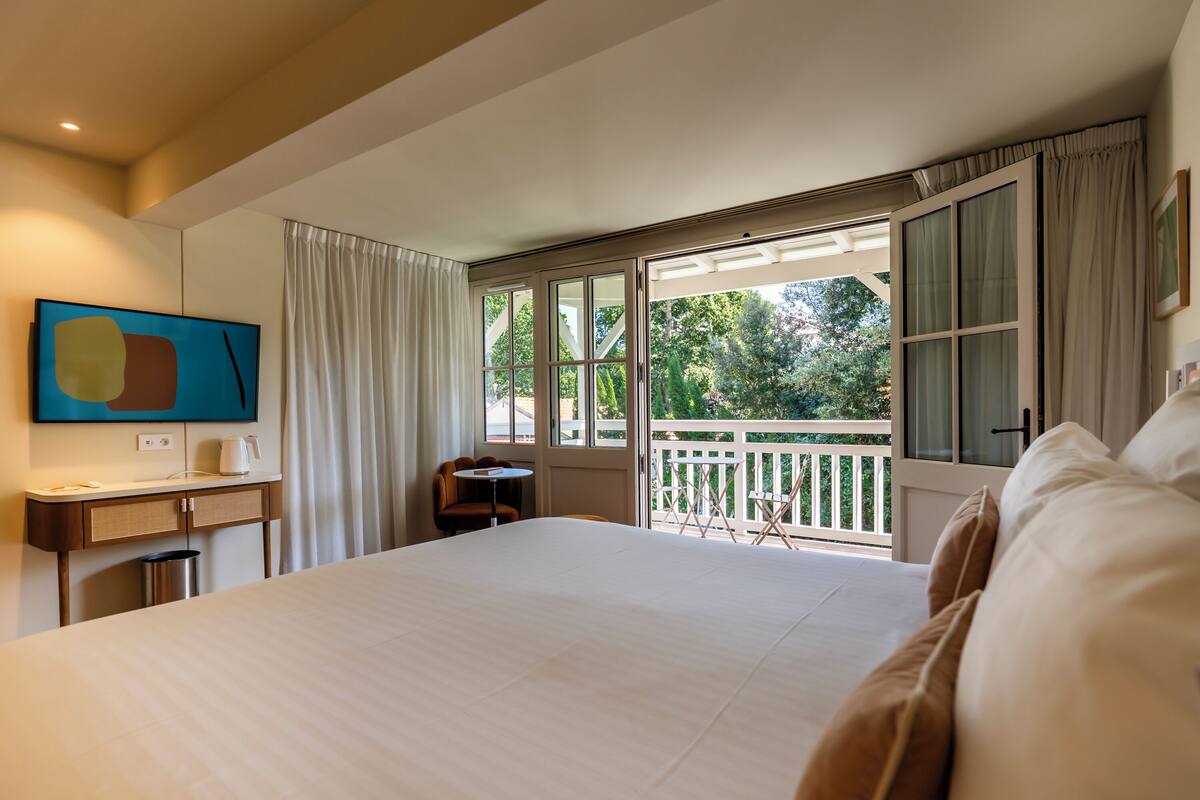 Habitación de hotel con una cama grande, un escritorio con silla, una mesita y vistas al exterior desde un balcón.