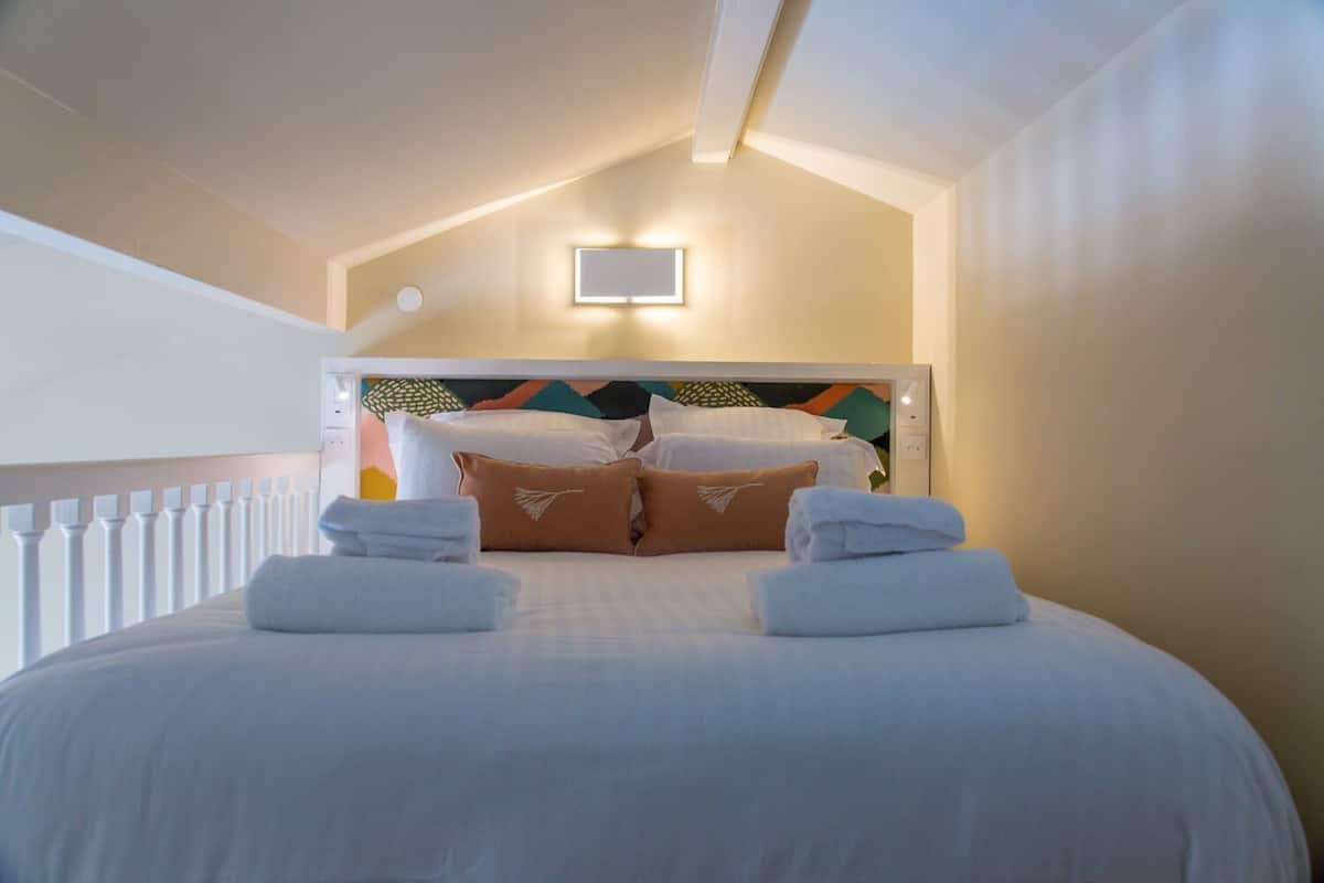 Habitación de hotel con cama, toallas blancas, cabecera con diseño geométrico, una luz pequeña montada en la pared y barandilla de balcón.