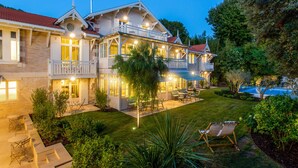 Une maison à deux étages avec un grand jardin à l’avant, un espace extérieur aménagé pour se détendre et une piscine.