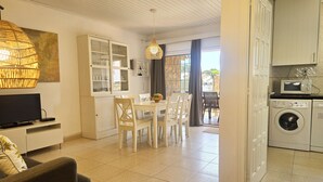 Un salon bien éclairé, équipé d’un téléviseur, d’un canapé, d’un coin repas et d’une cuisine avec un lave-linge.