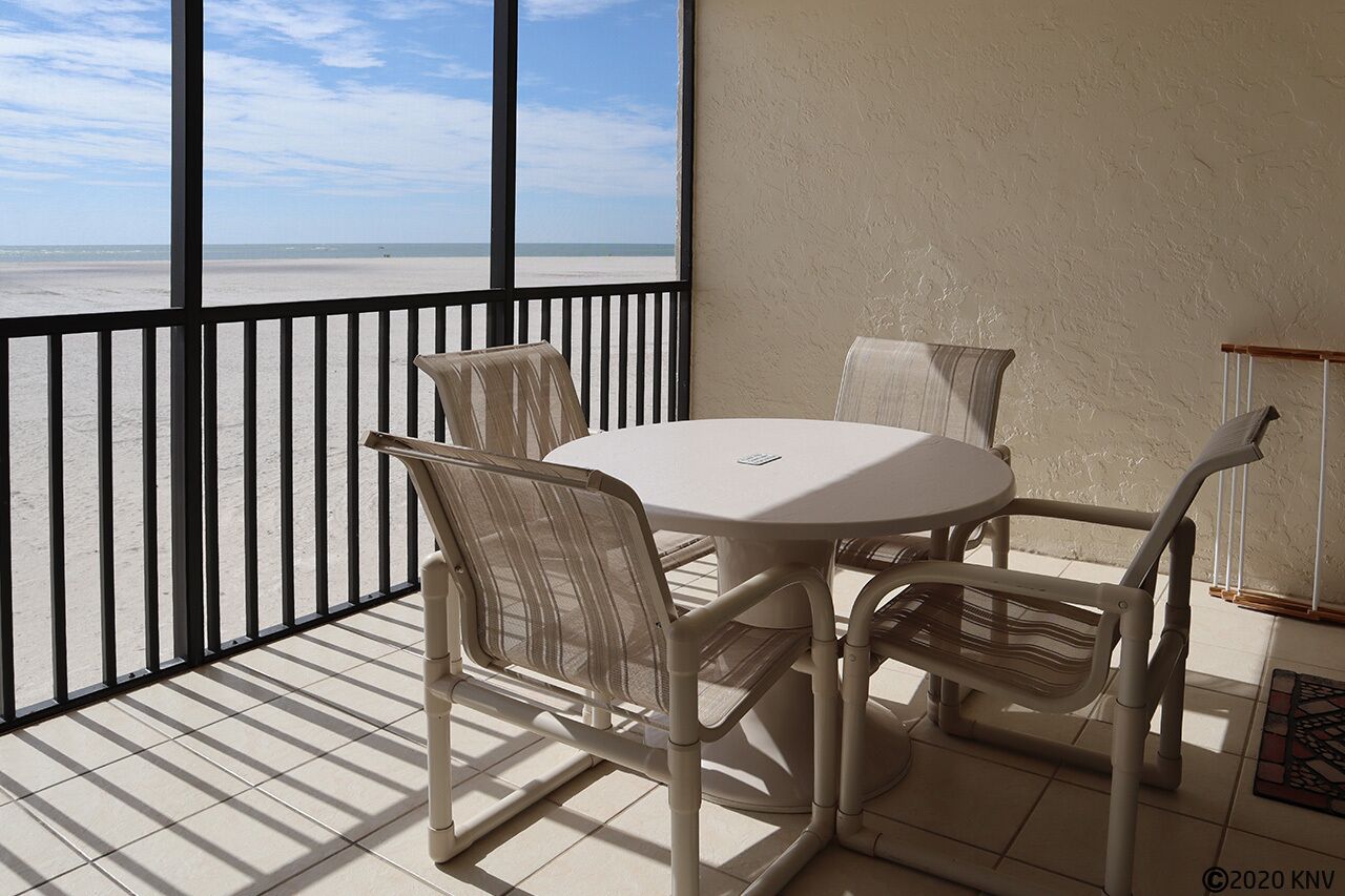 A balcony with a view of the beach, a round table with chairs, and a small side table.
