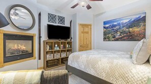 A bedroom with a fireplace, a television, a bookshelf, a bed, and a painting of a mountain landscape.