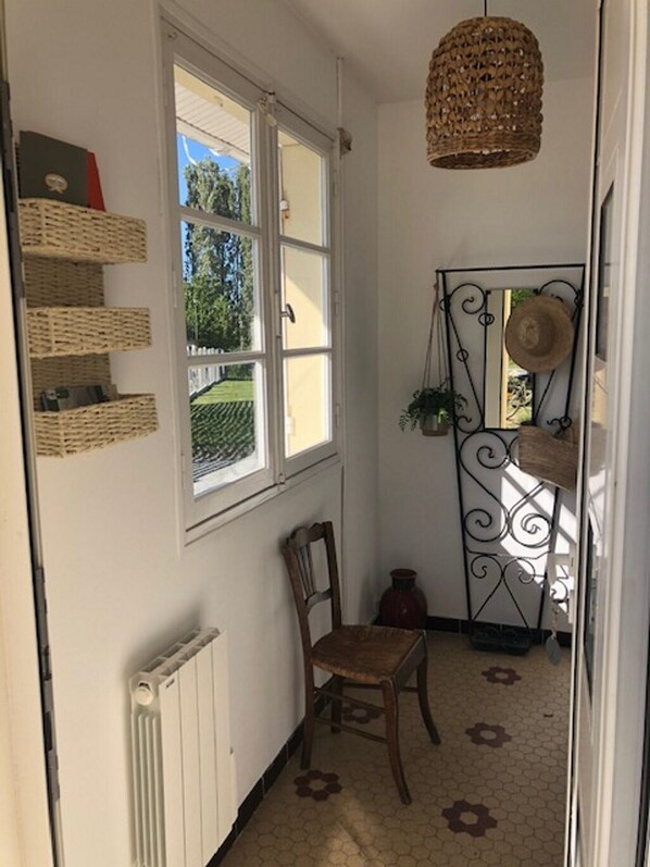 A narrow hallway with a radiator, a wooden chair, wicker baskets on the wall, a window with a view of greenery, and a decorative metal gate with a hat hanging on it.