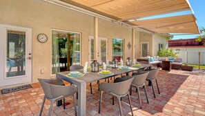 An outdoor patio with a dining table set for four, surrounded by wicker chairs, under a shaded area.