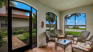 A covered patio with wicker furniture, a view of a landscaped garden, and a building with a tiled roof. - Beachy Condo in Artesia | Pool, Gym & Pickleball (Naples)