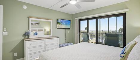 A bedroom with a large bed, a ceiling fan, a dresser, a television, and a view of the ocean through a sliding glass door.