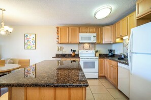 A kitchen with wooden cabinets, a granite countertop, a white stove, and a microwave. - Kamaole Sands #5-313 Idyllic Ocean Courtyard View, Across from Beach! A/C (Kihei)