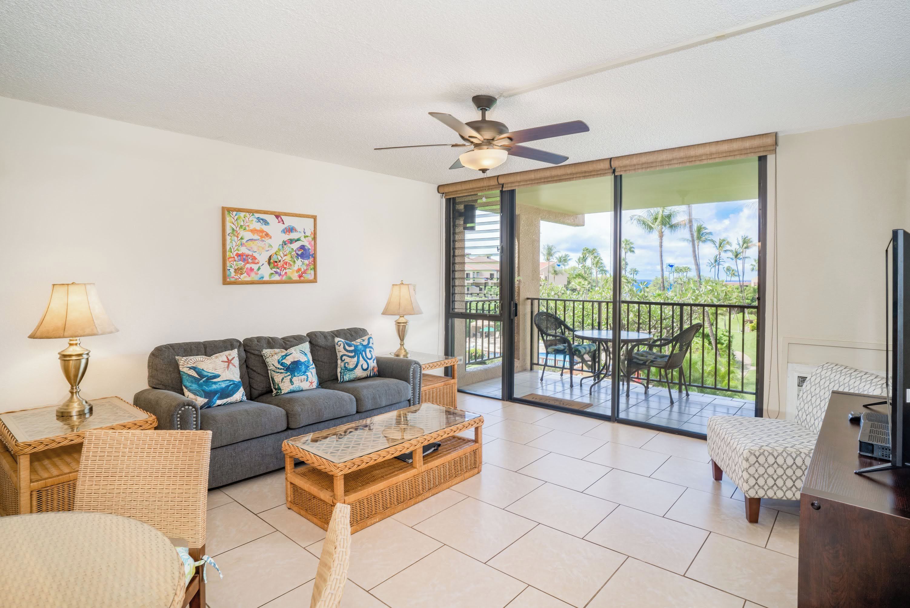 A spacious living room with a grey sofa, wicker chairs, a glass coffee table, and a ceiling fan. A balcony with outdoor furniture is visible through large sliding doors.
