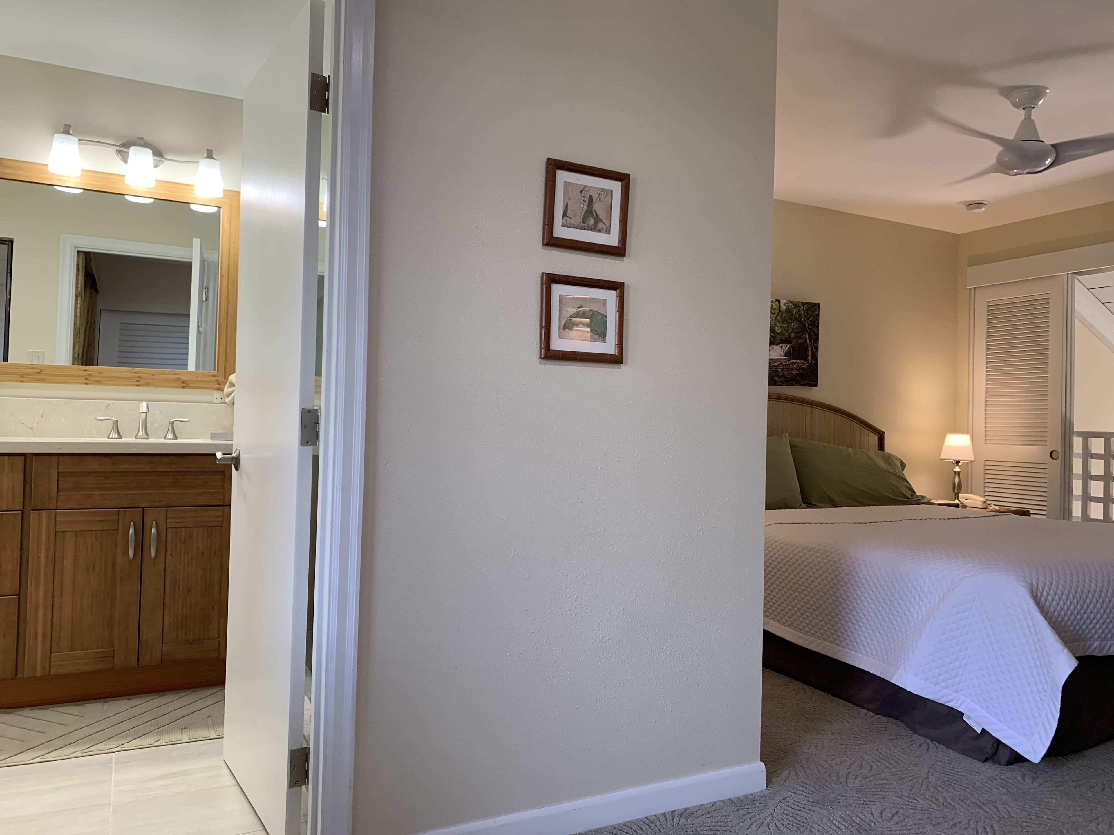 A bedroom with a bed, a nightstand, a lamp, a ceiling fan, and a door leading to a bathroom with a sink and mirror.