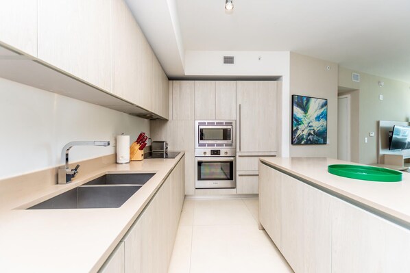 Une cuisine moderne avec des meubles en bois clair, des appareils en acier inoxydable, un grand îlot central doté d’un plateau vert et un tableau accroché au mur.