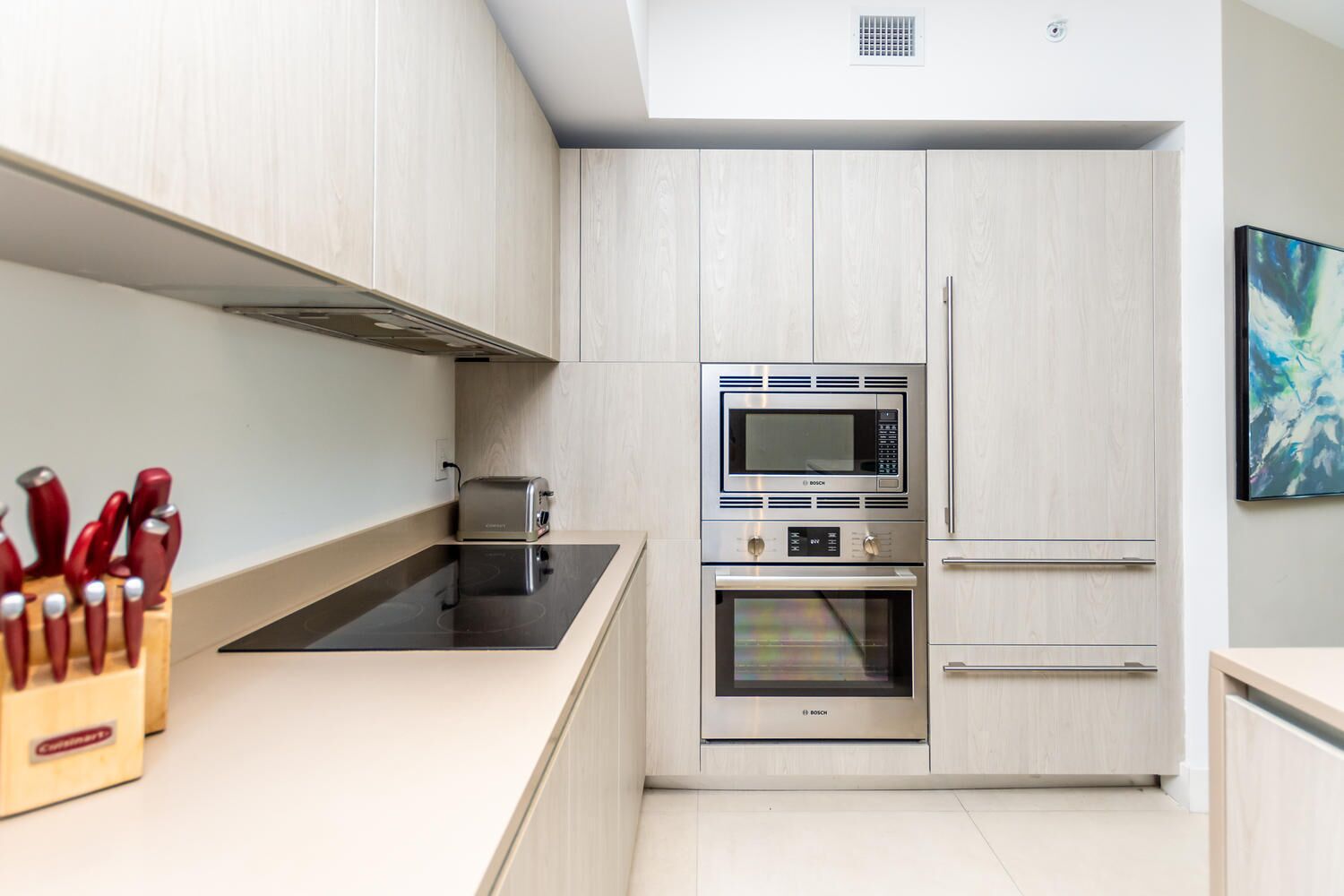 A modern kitchen with a Bosch microwave, toaster, and oven, light wood cabinets, and a countertop with a knife block.