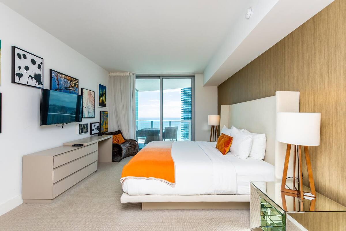 A modern hotel room with a large bed, a desk, a television, and a view of the city.