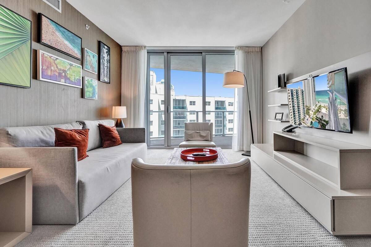 A modern hotel room with a sofa, a small table, a chair, a TV, and a large window with a view of buildings.