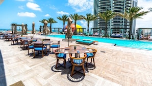 A poolside area with outdoor seating, a swimming pool, and a tall hotel building in the background. - Amazing Apartments at Lyfe Resort and Residences (Hollywood)